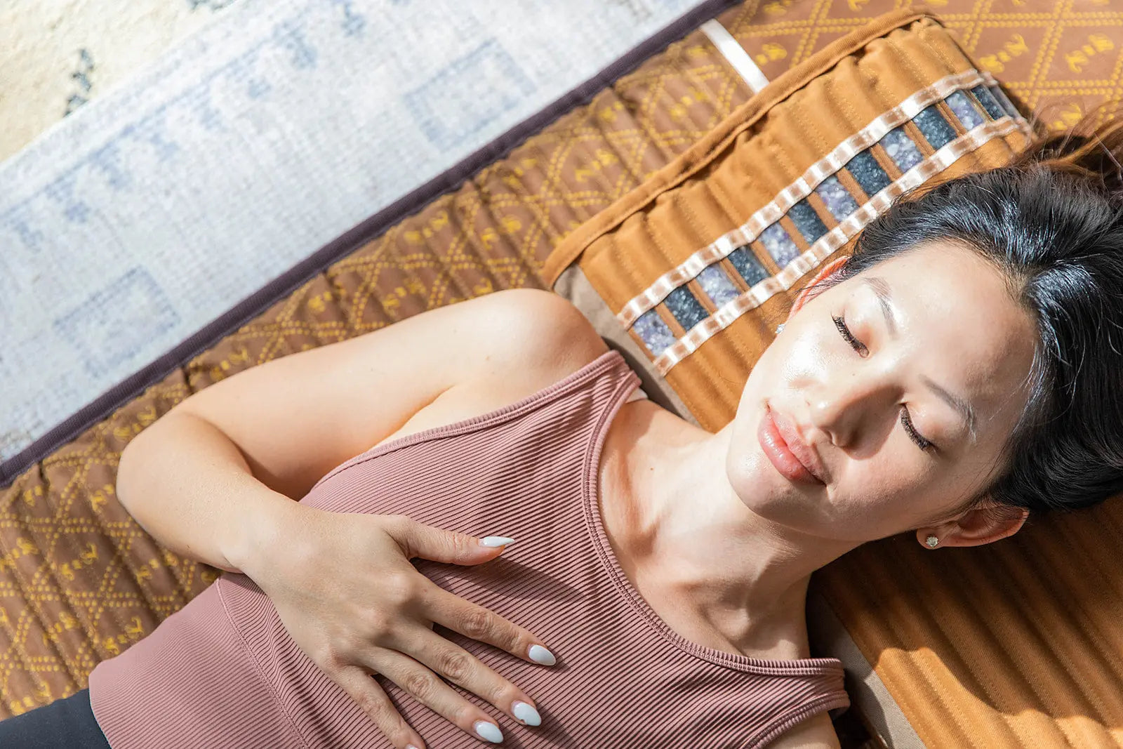Woman lying on a patterned brown mat with eyes closed, hand on chest, appearing relaxed