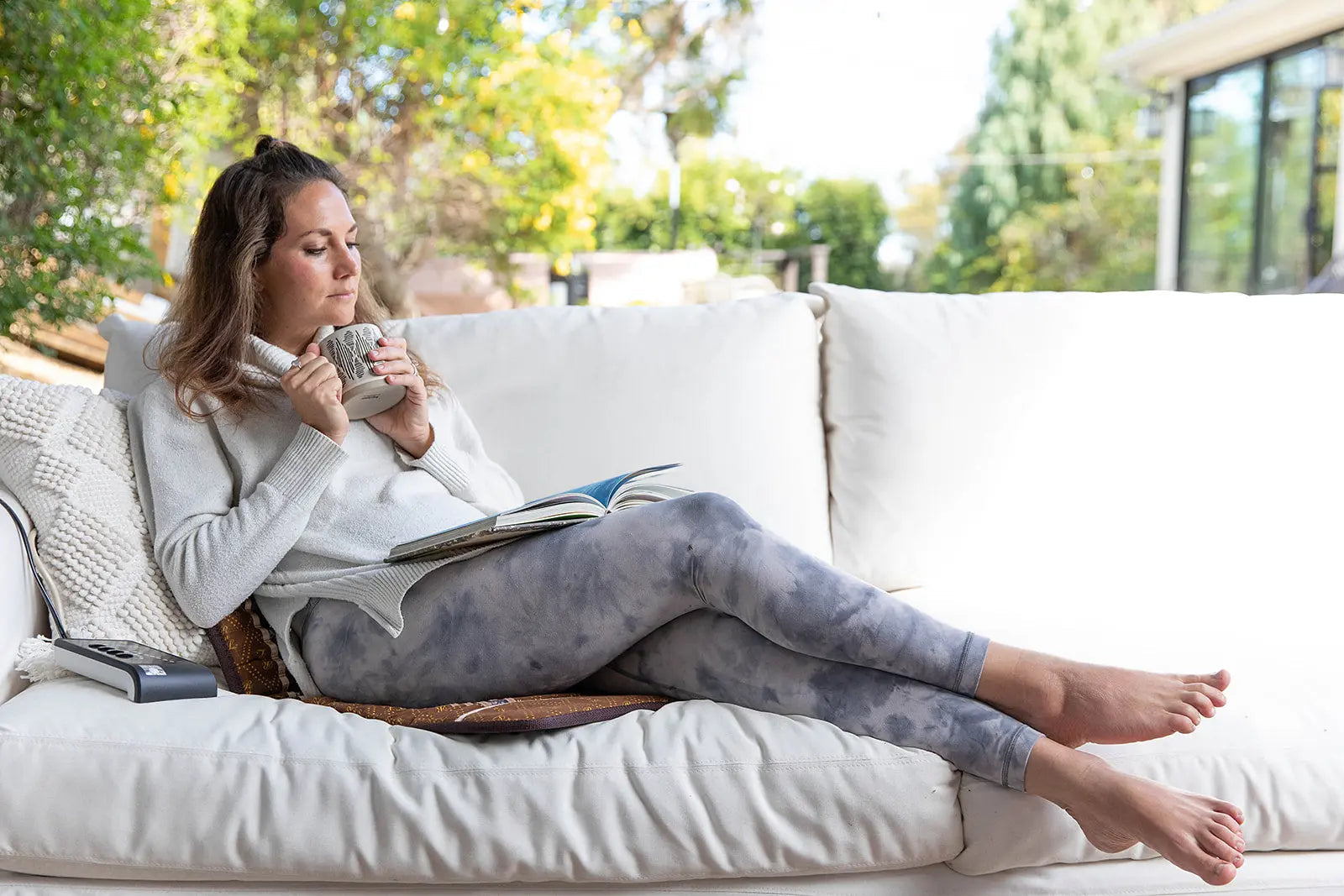Woman lounging on a white couch holding a mug and reading a book on a sunlit patio