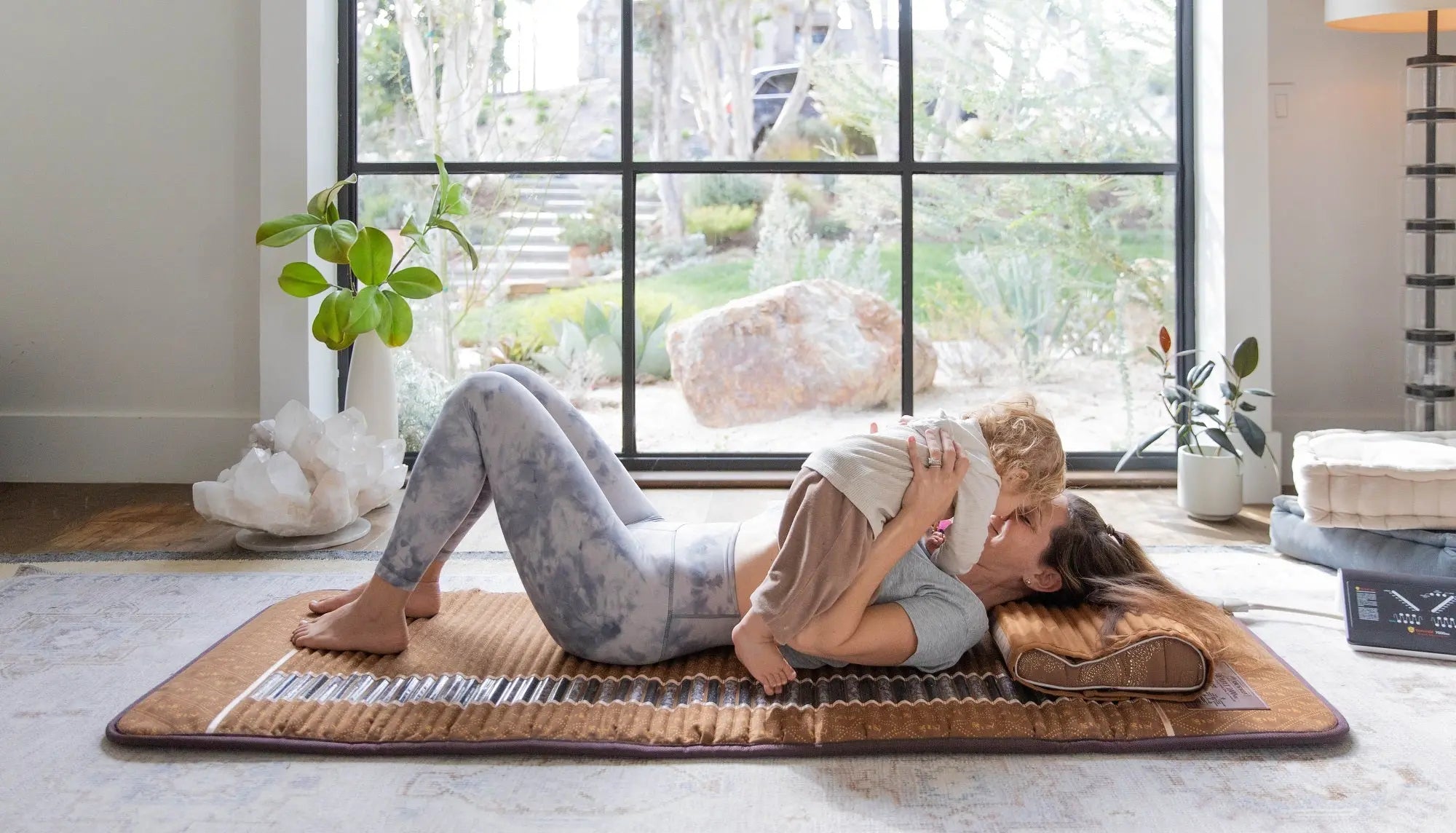 Mother lying on a brown BioMat holding her toddler in a bright living room by a large window with the controller nearby
