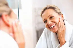 Woman smiling and touching her cheek while looking at herself in a bathroom mirror, appearing refreshed and youthful