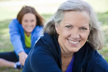 Older woman smiling while stretching on a mat in a sunny park with a younger woman assisting in the background