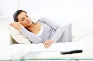 Woman napping on a white sofa, resting her head on a pillow in a bright living room setting