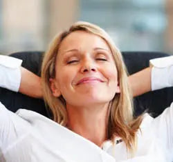 Woman reclining with hands behind her head, smiling with eyes closed in a bright, relaxed indoor setting