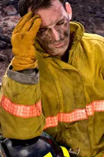 Firefighter in yellow turnout jacket sitting with hand to his forehead, looking exhausted with helmet at his side