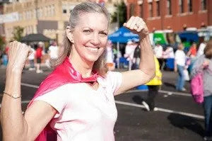 Smiling woman in pink cape flexing her arm at an outdoor cancer survivors celebration with people and booths in background