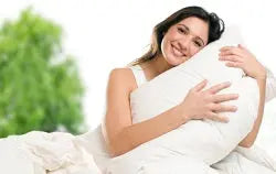 Smiling woman hugging a white pillow on a bright bed with blurred green plants visible through a window