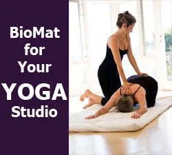 Two women doing assisted yoga stretches on a Biomat mattress with a purple banner reading “BioMat for Your YOGA Studio”