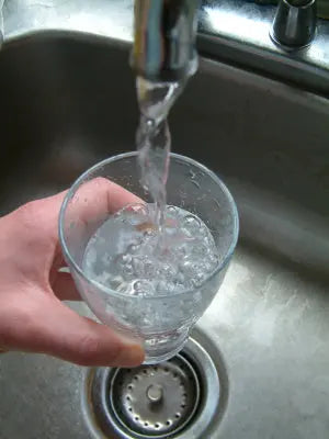 Hand holding a glass under a kitchen faucet as tap water fills the glass over a stainless steel sink