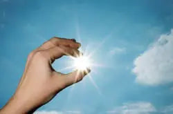Hand holding a bright sun-like light between fingers against a blue sky with a few clouds