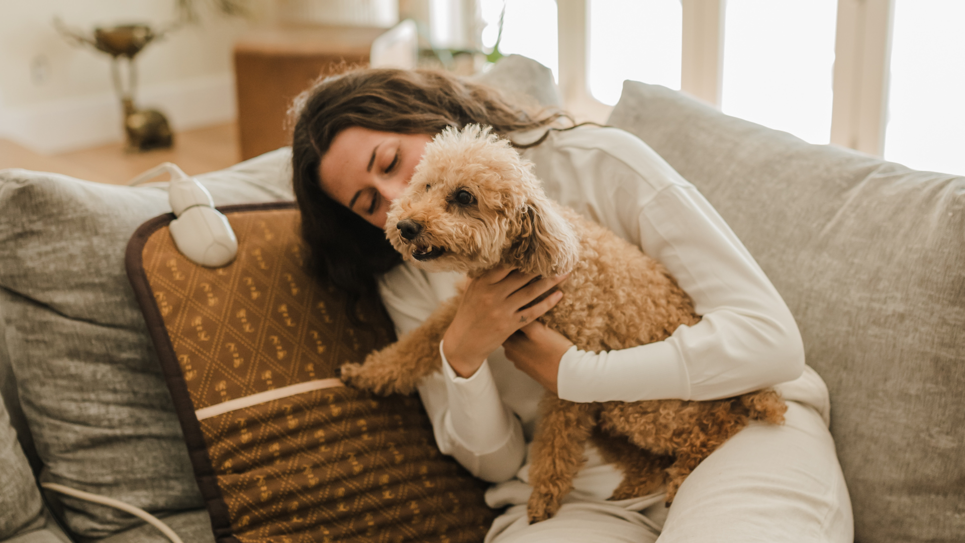 Woman hugging a small curly brown dog on a couch with a brown BioMat heated mat and visible controller