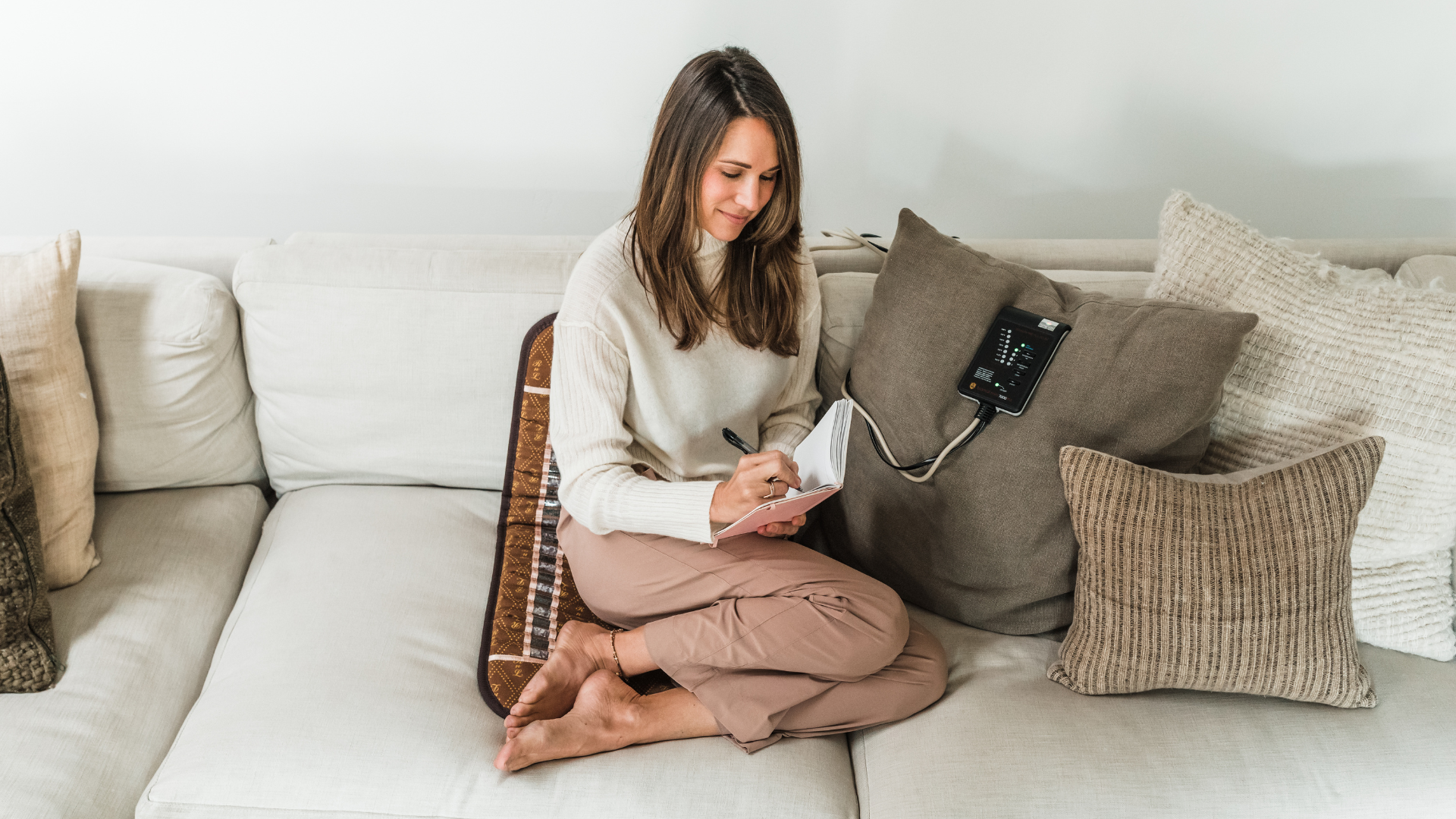 Woman sitting cross-legged on a couch writing in a journal while using a brown BioMat mat with its electronic controller resting on a pillow